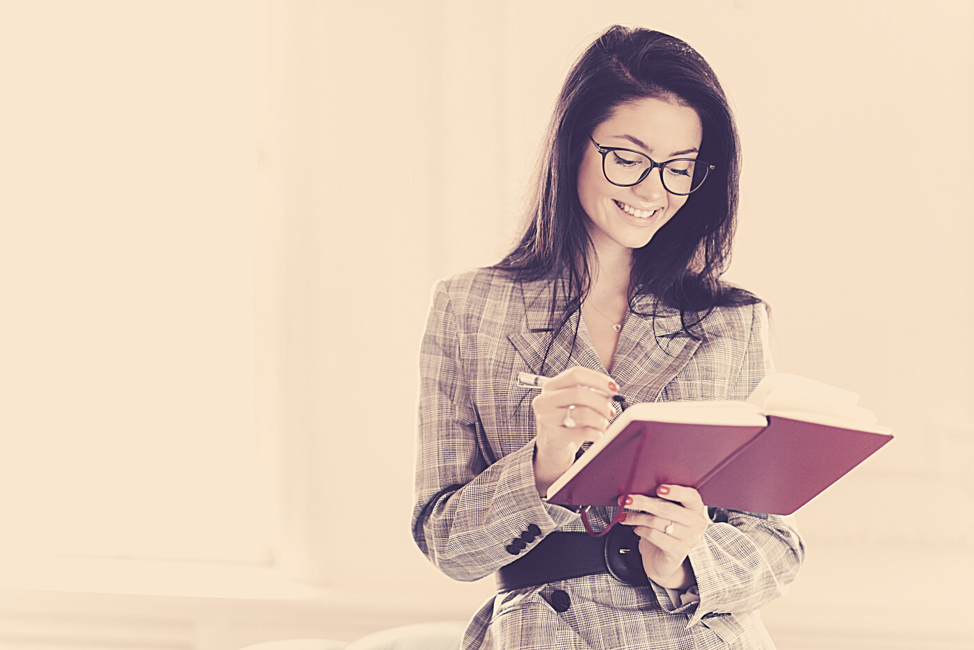 Horizontal Shot of Good Looking Woman Professor Writes down Text Notes in Notepad, Prepares for Lecture in University, Wears Transparent Glasses, Business Suit, Stands Indoor with Broad Smile